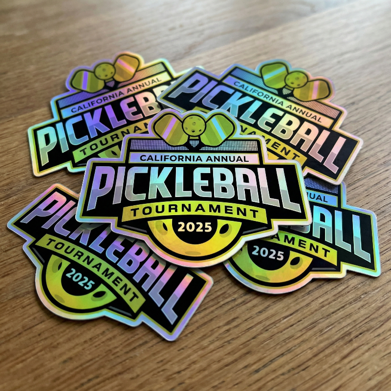 Holographic stickers for the California Annual Pickleball Tournament 2025 on a wooden surface