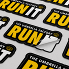 Custom Kiss-Cut Stickers with 'The Umbrella Factory Run' text on a gray background